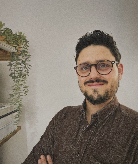 Portrait Homme Lunettes Brun, Chemise Casual Portrait d'un jeune homme brun portant des lunettes à monture écaille, vêtu d'une chemise boutonnée marron, posant les bras croisés près d'une étagère avec une plante suspendue.